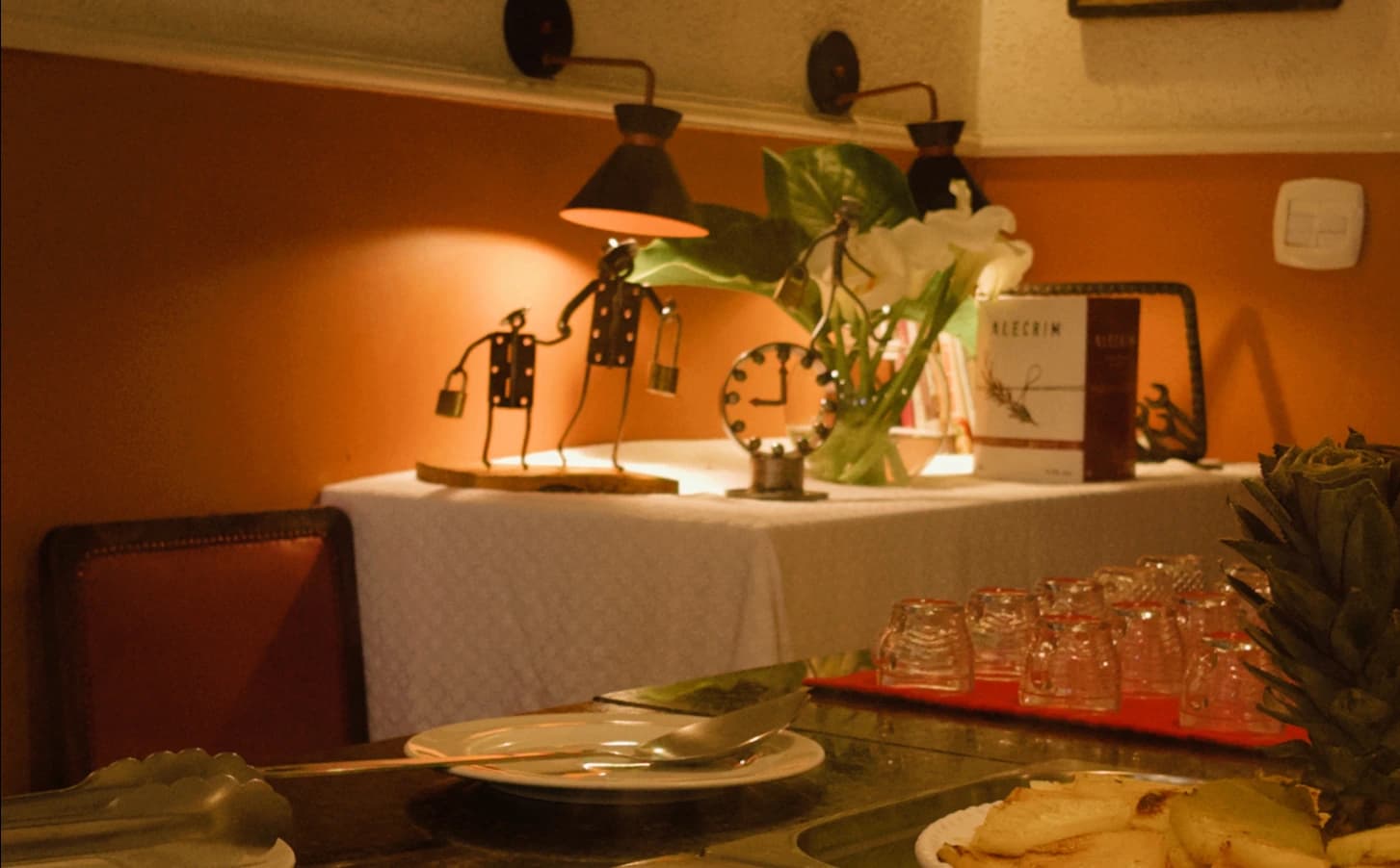 Salão do Bistrô Chef Lau à noite: mesa posta sob a luminária, com esculturas de sucata de Cláudio Silveira, relógio de engrenagens e antúrio — Muzambinho/MG